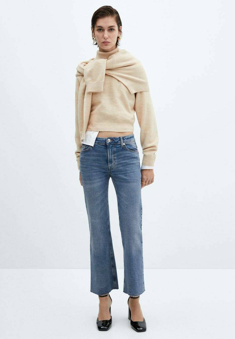 Vrouw met een beige trui aan en een andere trui over haar schouders gedrapeerd, blauwe cropped straight-leg jeans en zwarte hakken.