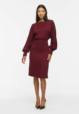 Una modelo posa sobre un fondo blanco liso con un vestido burdeos hasta la rodilla, de mangas voluminosas, y zapatos negros de tacón con punta afilada.