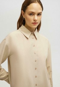 Chemise longue à manches beiges claires avec un col classique, fermeture à boutons à l'avant et texture de tissu douce, comportant des boutons assortis le long de la boutonnière.