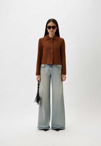 Bruine suède jas met een kraag, voorknopen en opgestikte zakken, gecombineerd met lichtblauwe jeans met wijde pijpen en een kleine zwarte tas.