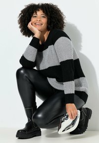 Schwarz-gestreifter Pullover in Schwarz und Grau, schwarze, gefälschte Lederleggings, klobige schwarze Stiefel mit Nieten und eine metallische Clutch-Tasche. Die gestrickte Textur ist sichtbar.