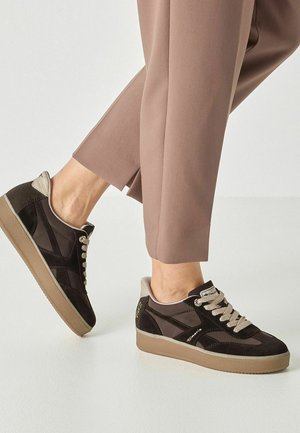 Persona que lleva zapatillas marrones y negras con suelas y cordones beige, combinadas con pantalones hasta el tobillo color topo sobre un fondo claro.