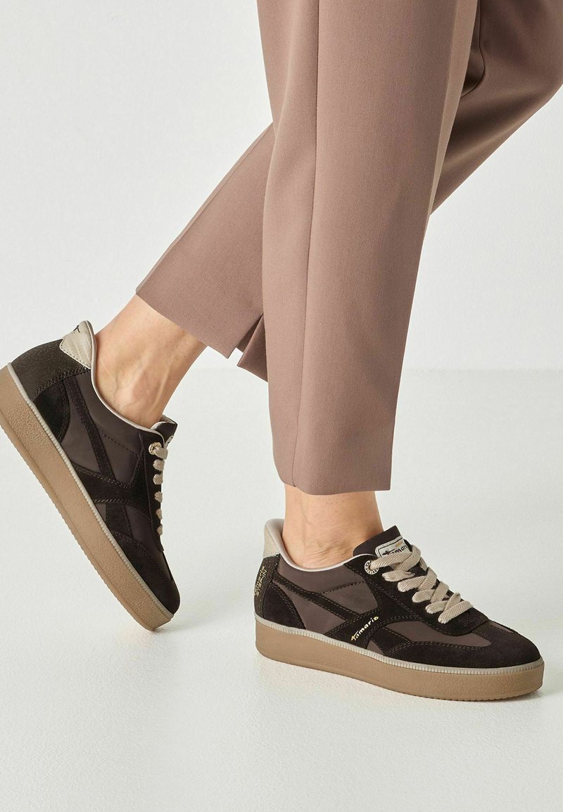 Personne portant des baskets marron et noires avec des semelles et lacets beiges, associées à un pantalon taupe longueur cheville sur un fond clair.