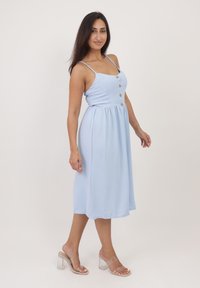 Vestido midi azul claro con tirantes finos, detalle de botones y cintura fruncida. Llevado con sandalias de tacón transparente sobre un fondo sencillo.