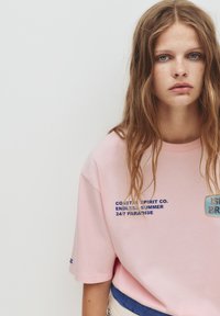 Lyserød oversized t-shirt lavet af bomuld med rund hals, prydet med mørkeblåt tekst og en rektangulær grafisk lap på brystet.