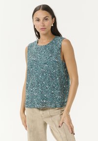 Haut sans manches en teal avec un motif abstrait ondulé. Tissu léger et ample avec un col rond. Assorti à un pantalon beige.