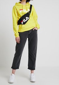 Žlutá mikina s bílým a červeným logem FILA, doplněná o černé džíny s ohrnutými nohavicemi a bílé tenisky. Malá černá kabelka přes rameno s logem FILA.