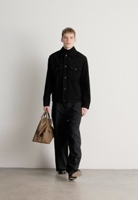 Melnas džinsa jaka ar priekšējām kabatām, melnas bikses un brūna tote soma. Modelis pastaigājas uz neitrālas krāsas fona. Gluda tekstūra un tīras līnijas.