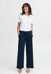Pantaloni a gamba larga blu navy con una texture liscia, dotati di tasche laterali e un singolo bottone frontale. Abbinati a una t-shirt bianca a maniche corte semplice.