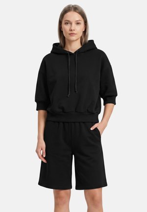 Kvinde står iført en sort kortærmet hættetrøje og matchende sorte knælange shorts med den ene hånd i lommen mod en hvid baggrund.