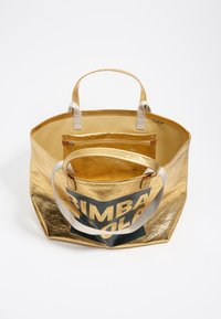 Złota metaliczna torba na ramię z materiałowymi uchwytami, posiada jednolitą ciemnozieloną panel z logo oraz wewnętrzną kieszeń na zamek. Wnętrze wyłożone jest złotym materiałem.