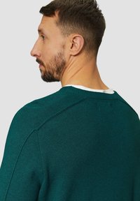Türkiser Rundhals-Pullover aus strukturiertem Strickstoff, mit Raglanärmeln und einem kontrastierenden weißen Kragen. Die Rückansicht zeigt Detailnähte an den Schulterensäumen.