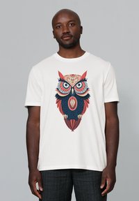 Weißes Baumwoll-T-Shirt mit einem farbenfrohen Eulendesign, das detaillierte Elemente in Rot, Blau und Schwarz aufweist. Kurze Ärmel und runder Halsausschnitt.