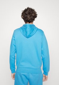 Sudadera con capucha de color azul brillante, con una textura suave, puños y cinturilla acanalados. Vista desde la parte trasera, destacando la capucha y las costuras.