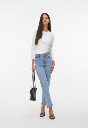 Haut à manches longues blanc, jeans taille haute bleu clair, sac noir et sandales à lanières noires, sur un fond blanc uni.