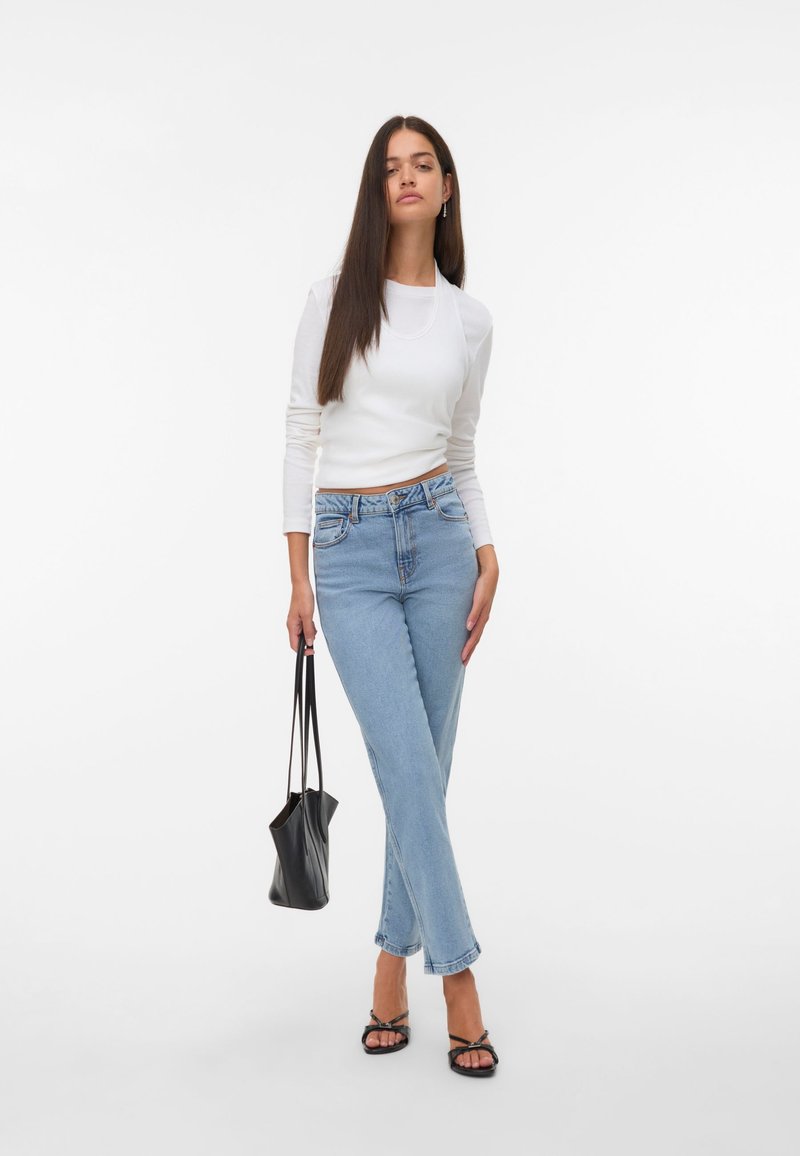 Bela dolga majica z dolgimi rokavi, svetlo modri visokorasli jeans, črna torba in črne strappy sandale, na enobarvni beli ozadju.
