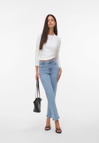 Balta ilgomis rankovėmis palaidinė, šviesiai mėlynos kelnės su aukštu liemeniu, juoda rankinė ir juodos sandalai su dirželiais, prieš baltą fono.