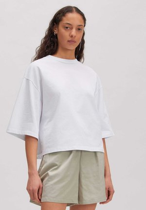 Jonge vrouw met lang krullend haar, die een wit oversized t-shirt en beige korte broek draagt, staand tegen een effen lichte achtergrond.