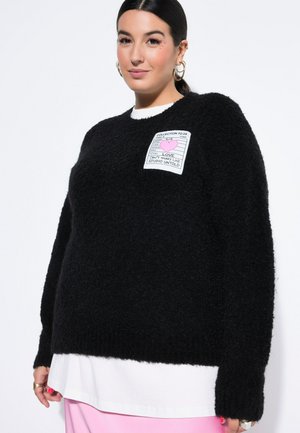 Donna con capelli scuri indossa un maglione nero peloso con una toppa bianca con la scritta "LOVE CAN'T HURRY LOVE" sopra una camicia bianca e una gonna rosa, sorridendo leggermente.