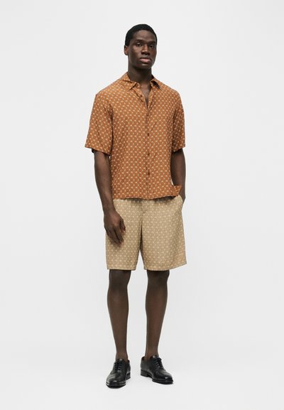 Άνδρας με καφέ πουκάμισο με κοντά μανίκια και σχέδιο, μπεζ σορτς με σχέδιο και μαύρα επίσημα παπούτσια, στέκεται μπροστά σε απλό φόντο.