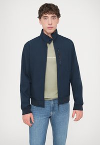 Μπουφάν μπόμερ σε ναυτικό μπλε με όρθιο γιακά, φερμουάρ μπροστά και ριμπ μανσέτες, φορεμένο πάνω από ανοιχτό πράσινο πουκάμισο και συνδυασμένο με μπλε τζιν.