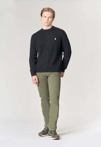 Maglione grigio scuro a treccia, pantaloni verde oliva slim fit e sneakers verdi; design semplice con polsini a costine e piccolo logo sul maglione.