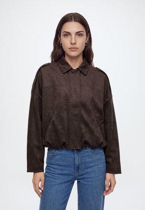 Femme aux longs cheveux bruns portant une veste marron foncé et un jean bleu, debout devant un fond clair uni avec une expression neutre.