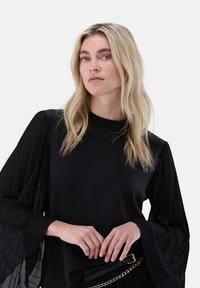 Blonde Frau mit welligem Haar trägt eine schwarze Bluse mit durchscheinenden, plissierten langen Ärmeln, schwarzen Gürtel mit goldener Kette und posiert vor einem schlichten Hintergrund.