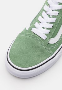 Gröna mocka sneakers med en vit sidrand, vita snören, svarta detaljer och en texturerad gummisula. Nära vy av tåområdet.