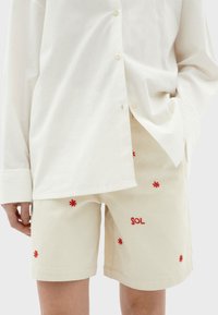 Beige katoenen shorts met rode bloemenborduring en het woord "SOL." Gecombineerd met een losse witte blouse, met zichtbare knopen en manchetten.