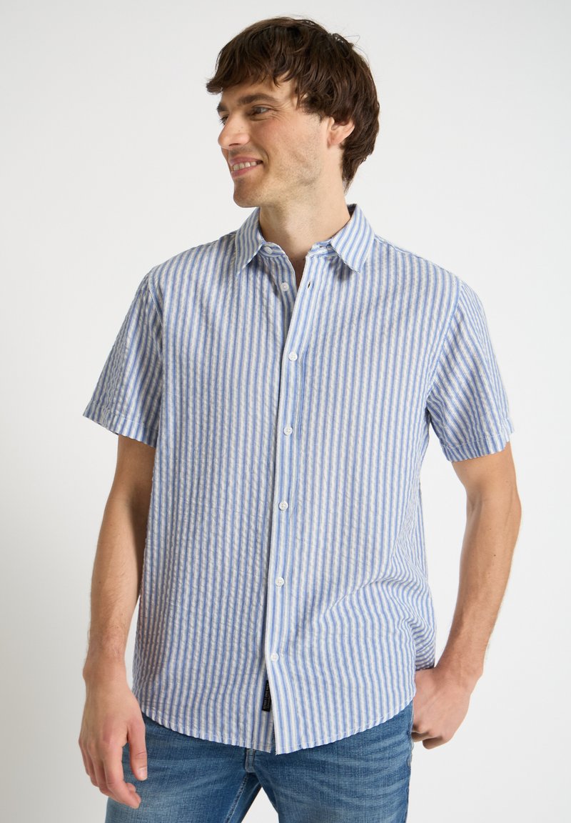 Homme portant une chemise à manches courtes rayée verticale bleue et blanche avec col, souriant et regardant vers la gauche.