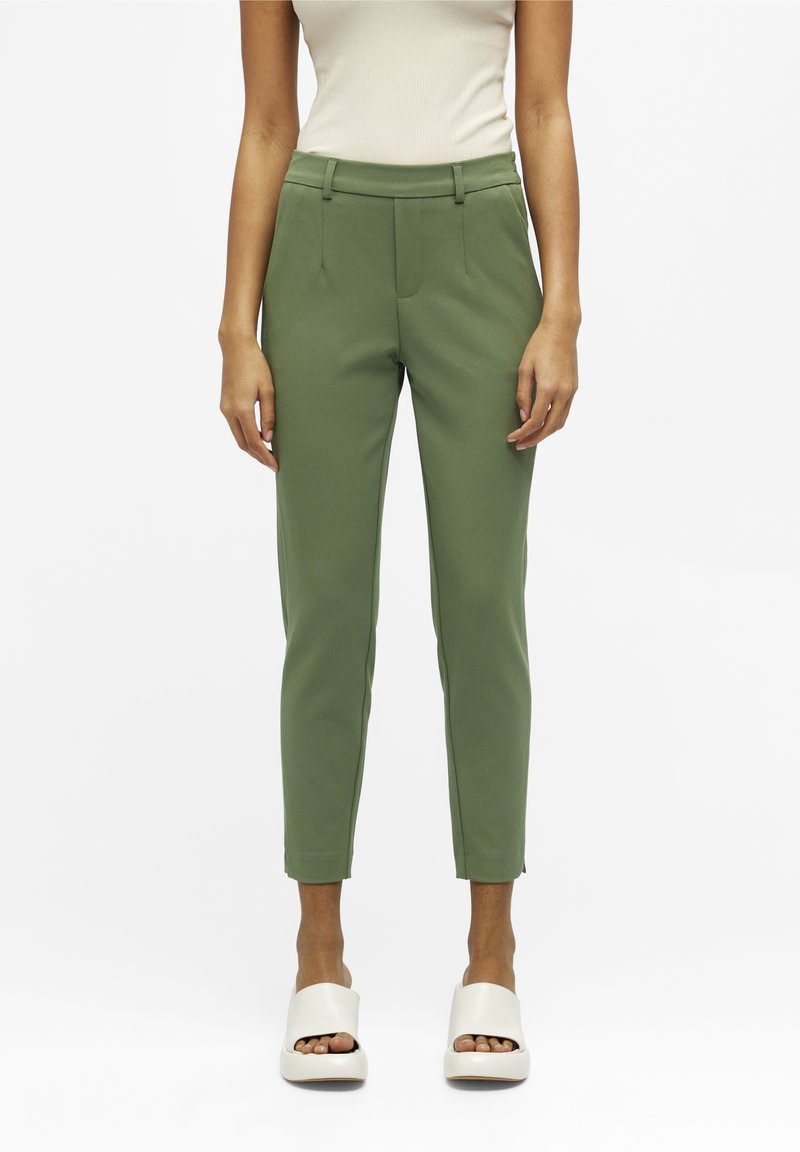 Pantalon vert en tissu extensible, coupe ajustée avec poches latérales, design à jambes fines et texture lisse. Porté avec des chaussures blanches à bouts ouverts.