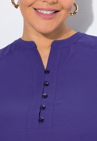 Maglietta viola con collo rotondo e cinque bottini sul davanti. Il tessuto sembra liscio, con dettagli di cucitura delicati sulle spalle.