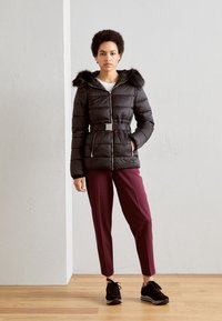 Černá péřová bunda s kapucí lemovanou kožešinou, se zipem vpředu, páskem v pase a dvěma bočními kapsami, v kombinaci s vínově červenými kalhotami a černými teniskami.