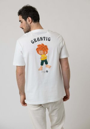 Mann in weißem T-Shirt mit Cartoonfigur und dem Wort "GRANTIG" darüber auf dem Rücken, trägt hellbeige Hose.