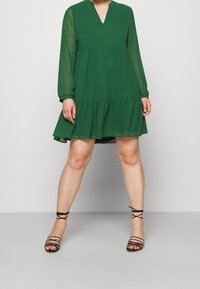Zielona sukienka z dekoltem w serek, długimi przezroczystymi rękawami i falbanowym dołem. Zestawiona z czarnymi sandałami na paski. Gładka faktura tkaniny.