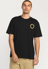 T-shirt en coton noir à manches courtes, col rond, avec un imprimé graphique jaune sur la poitrine gauche, présentant un design de logo circulaire.