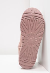 Suela de una bota UGG rosa con un patrón de tracción texturizado en forma de ráfaga solar, apoyada contra una superficie blanca en la esquina.