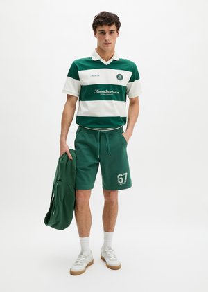 Jeune homme portant un polo rayé vert et blanc avec le texte "Scandinavian", short vert avec le numéro 67, tenant une veste assortie, baskets blanches.