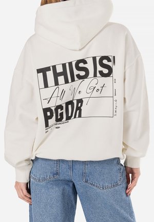 Persona con una sudadera blanca con capucha y un gran texto negro "ESTO ES PGDR Todo lo que tenemos" en la espalda, combinada con vaqueros azules, vista desde atrás.