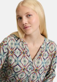 Multicolored patterned blouse with geometric designs in blue, pink, and yellow. Features a partial button placket and textured neckline detail.