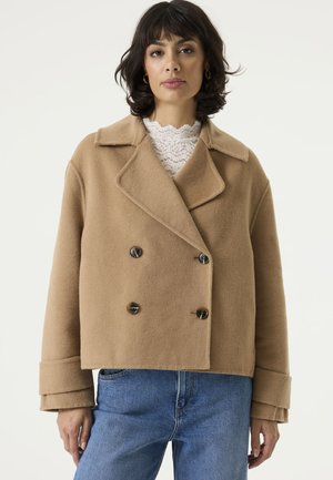 Woman with dark hair wearing a tan double-breasted wool coat, white lace top, and blue jeans, standing against a plain background.