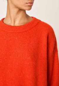 Femme portant un pull rouge vif en tissu duveteux avec un col rond côtelé, visible du cou au milieu du torse sur un fond uni.