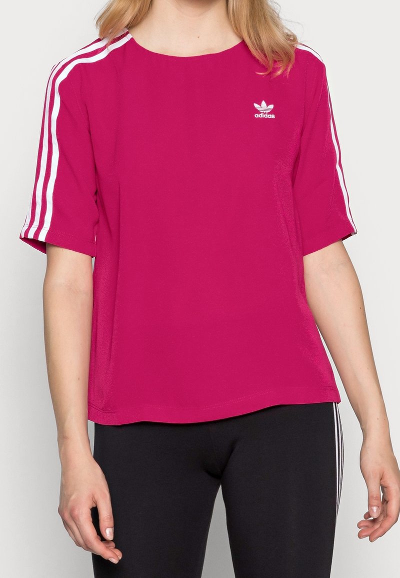 Růžový sportovní top s krátkým rukávem, se třemi bílými pruhy na ramenou a malým bílým logem na hrudi.