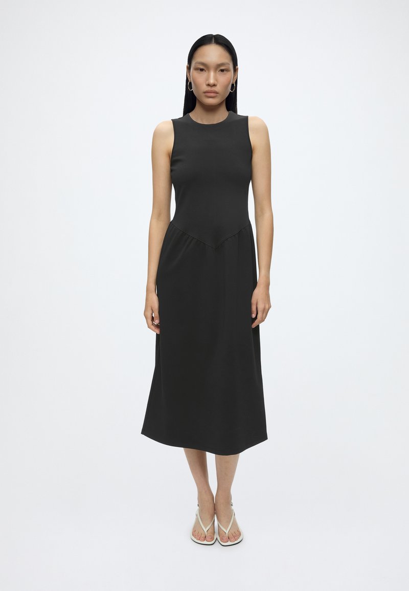 Femme aux longs cheveux noirs portant des boucles d'oreilles créoles et une robe midi noire sans manches avec des sandales blanches, debout devant un fond blanc uni.