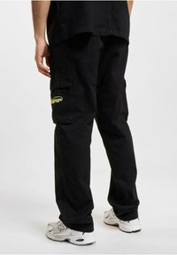 Black cargo pants with multiple pockets, featuring yellow text detail on the left pocket. Made of durable fabric, relaxed fit, straight leg design.