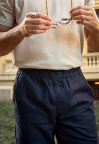 Camiseta de manga corta color beige claro, pantalones cortos de corte relajado en navy con cinturilla elástica, manos sosteniendo gafas de sol redondas con acentos dorados.