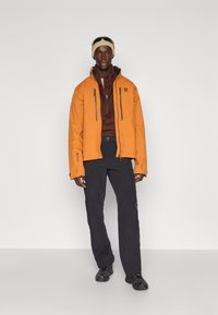 Oranje waterdichte jas met hoge kraag, zwarte ritsen en twee voorzakken, gecombineerd met donkere broek en zwarte schoenen.