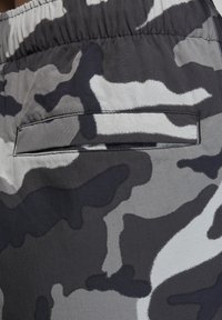 Kamouflagemönstrad tyg i grå och svarta nyanser; inkluderar en ficka med patchdetalj. Slät textur med elastisk midja.