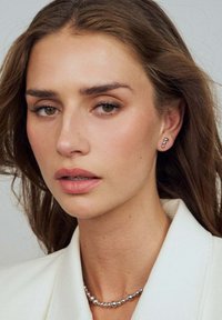 Silverörhängen med en böjd design, bärs tillsammans med ett pärlhalsband. Modellen har mellanbrunt hår, neutral makeup och en vit kavaj.